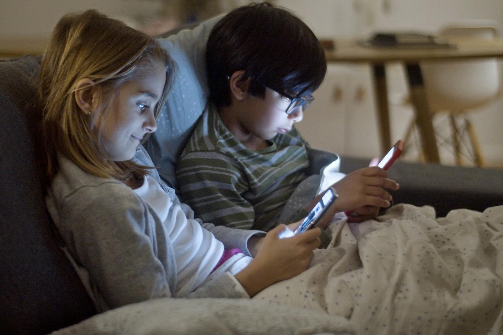 Two children using smartphones at night, indoors, under blankets.