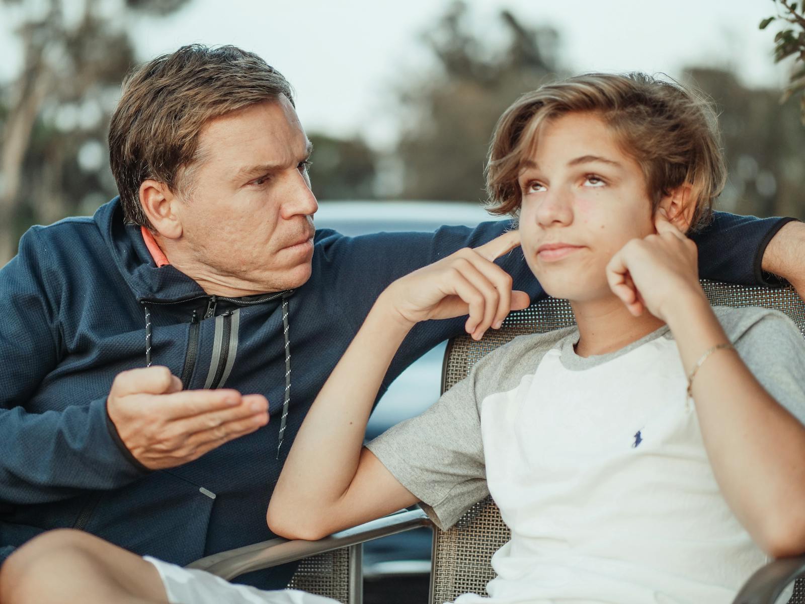 Father expressing frustration while teenage son shows defiance, seated outdoors. Get Your Child to Listen Without Yelling
